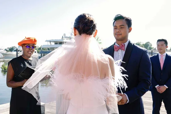A wedding officiant in a black dress and orange beret officiating a wedding ceremony for a couple on a dock.