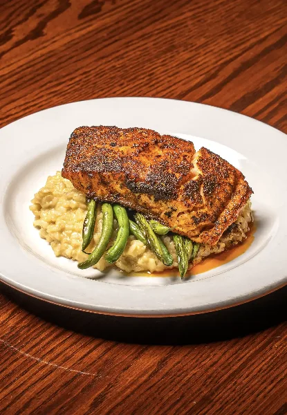 A plated restaurant dish featuring a seared protein over grits with green beans on a white plate on a wooden table.