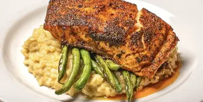 A plated restaurant dish featuring a seared protein over grits with green beans on a white plate on a wooden table.