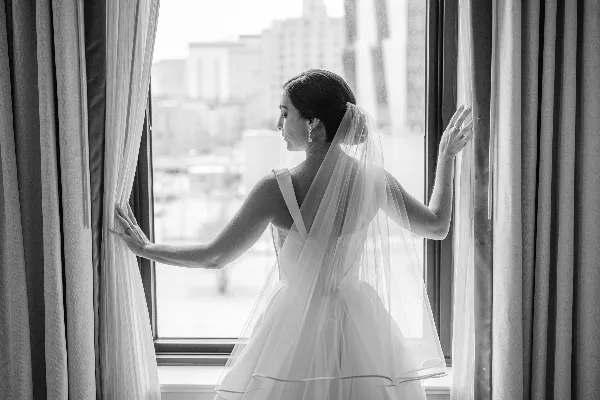 A black and white photograph of a bride in a wedding dress and veil standing by a window, looking out at a city skyline.