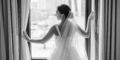 A black and white photograph of a bride in a wedding dress and veil standing by a window, looking out at a city skyline.