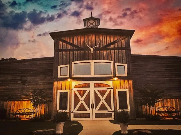 Exterior view of a rustic wooden event venue building with a mounted antler on the facade and white double doors at dusk.