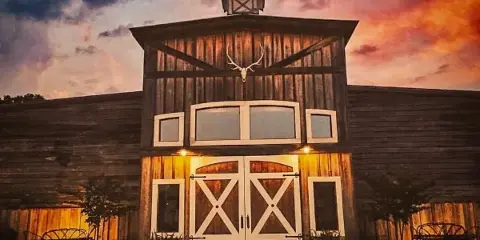 Exterior view of a rustic wooden event venue building with a mounted antler on the facade and white double doors at dusk.