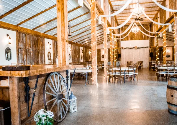 Interior of a rustic barn wedding venue featuring a wooden bar, string lights, and reception tables.