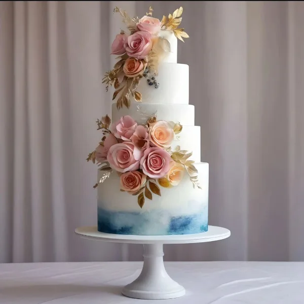 A five-tier white wedding cake decorated with pink roses and gold leaves, featuring a blue watercolor base, displayed on a white pedestal stand.