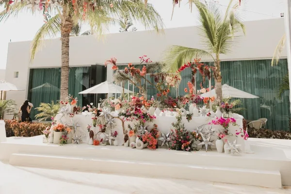 A professionally decorated wedding event space featuring a long table with elaborate floral arrangements, star-shaped decor, and a white backdrop in a tropical setting.