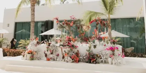A professionally decorated wedding event space featuring a long table with elaborate floral arrangements, star-shaped decor, and a white backdrop in a tropical setting.
