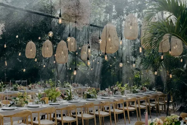 An outdoor event table set with white linens, wooden chairs, floral arrangements, and hanging woven pendant lights.