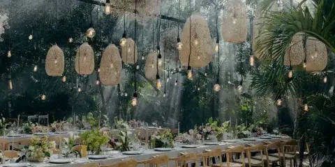 An outdoor event table set with white linens, wooden chairs, floral arrangements, and hanging woven pendant lights.