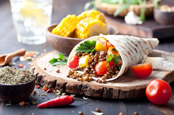 A close-up photograph of a food wrap filled with vegetables and lentils, served on a wooden board with fresh corn and tomatoes.