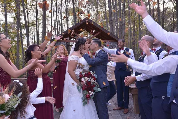 A bride and groom share a kiss during an outdoor wedding ceremony while guests and the wedding party celebrate around them.