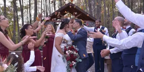 A bride and groom share a kiss during an outdoor wedding ceremony while guests and the wedding party celebrate around them.