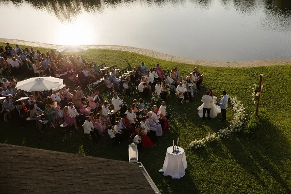 An outdoor wedding ceremony with guests seated on a lawn next to a lake at sunset.