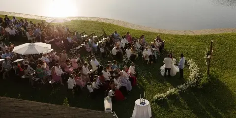 An outdoor wedding ceremony with guests seated on a lawn next to a lake at sunset.
