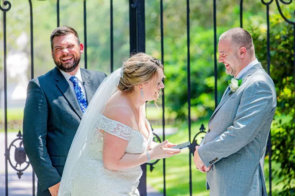A bride and groom smiling and exchanging rings during an outdoor wedding ceremony.
