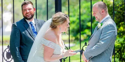 A bride and groom smiling and exchanging rings during an outdoor wedding ceremony.
