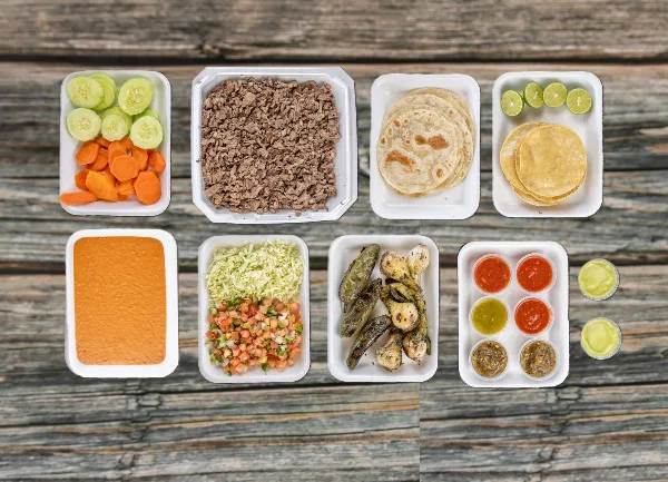 A catering spread of carne asada, tortillas, salsas, and vegetables in white takeout containers on a wooden table.