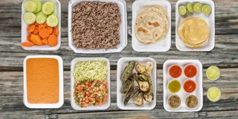 A catering spread of carne asada, tortillas, salsas, and vegetables in white takeout containers on a wooden table.