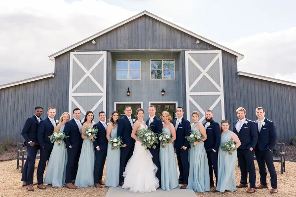 A wedding party consisting of a bride, groom, and attendants posing in front of a rustic gray barn.