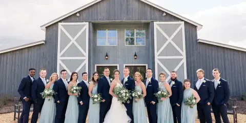 A wedding party consisting of a bride, groom, and attendants posing in front of a rustic gray barn.