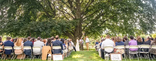 An outdoor wedding ceremony with guests seated in rows facing a couple under a large tree.