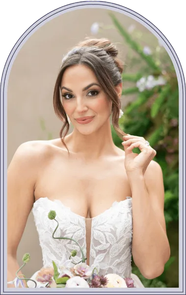 A bride in a white lace wedding dress with a styled updo and full makeup, holding a bouquet.