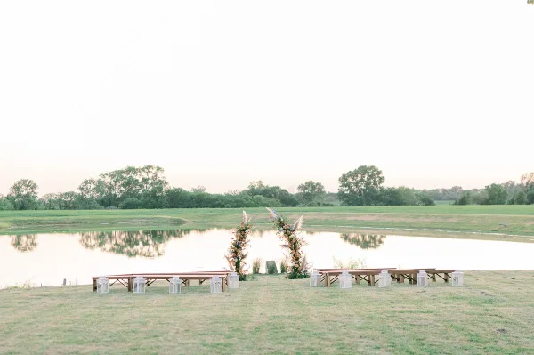 Outdoor wedding ceremony setup with a floral arch and wooden benches overlooking a lake and green fields.