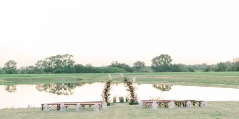Outdoor wedding ceremony setup with a floral arch and wooden benches overlooking a lake and green fields.