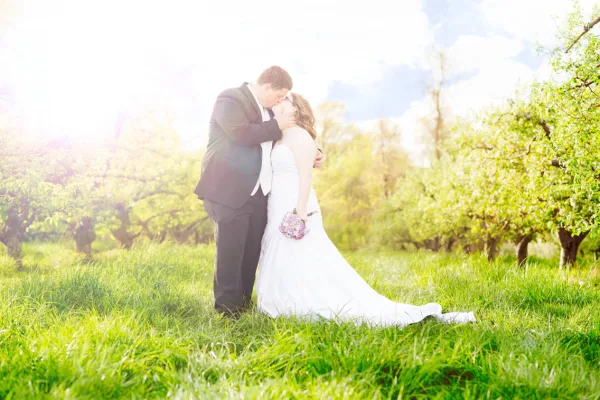 A bride and groom embracing in a grassy field with trees in the background.