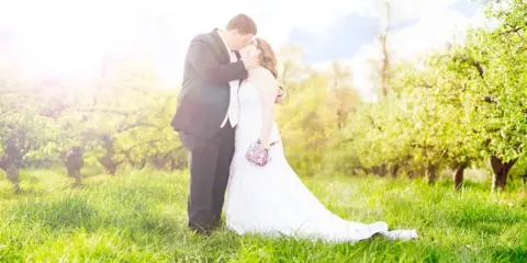 A bride and groom embracing in a grassy field with trees in the background.