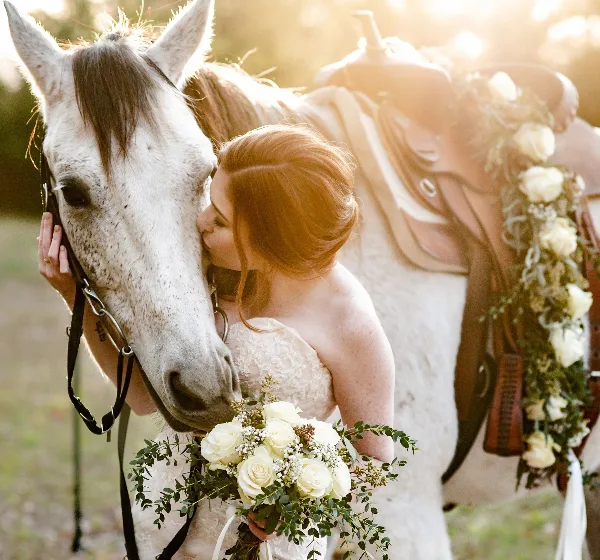A bride in a white lace wedding dress holding a bouquet of white roses and eucalyptus, kissing the nose of a white horse with a saddle decorated with flowers.