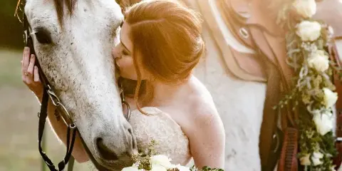 A bride in a white lace wedding dress holding a bouquet of white roses and eucalyptus, kissing the nose of a white horse with a saddle decorated with flowers.