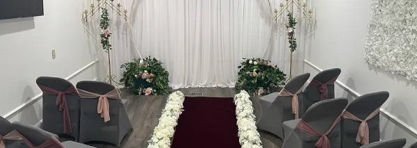 A wedding ceremony setup featuring a red carpet aisle lined with white flowers, grey chairs with sashes, and floral arrangements against a white backdrop.