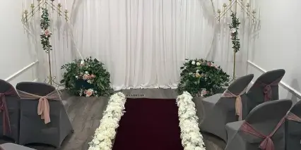 A wedding ceremony setup featuring a red carpet aisle lined with white flowers, grey chairs with sashes, and floral arrangements against a white backdrop.