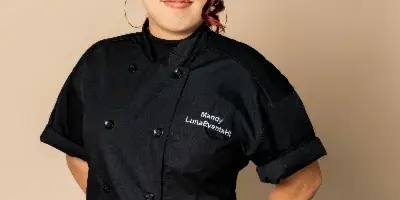 Chef in a black Luna Events chef coat standing behind a table with a tomato, knife, and cutting board.