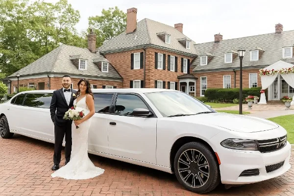 A bride and groom posing in front of a white stretch limousine at a brick building venue.