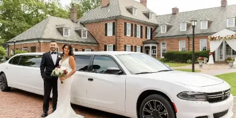 A bride and groom posing in front of a white stretch limousine at a brick building venue.