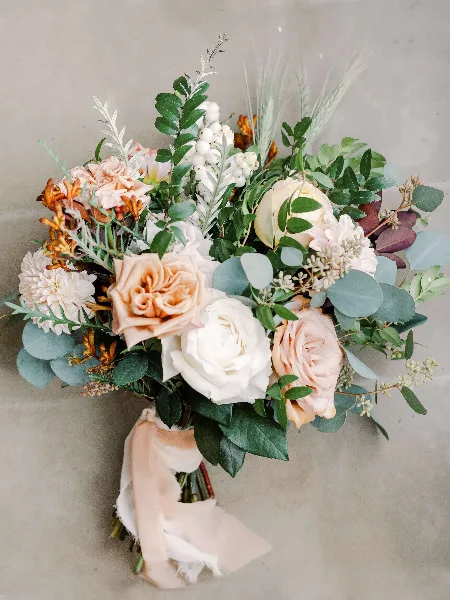 A close-up of a wedding bouquet featuring peach and white roses, eucalyptus leaves, and dried grasses tied with a ribbon.