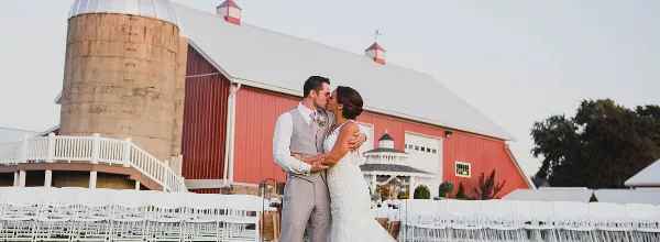A bride and groom kissing in front of a large red barn and silo with white chairs set up for a wedding.
