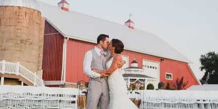 A bride and groom kissing in front of a large red barn and silo with white chairs set up for a wedding.
