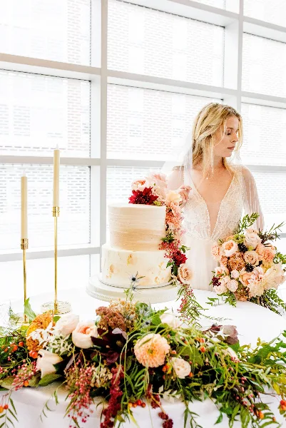 A bride holding a bouquet of pink and peach roses stands next to a wedding cake adorned with cascading floral arrangements and candles.