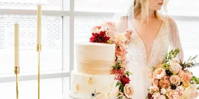 A bride holding a bouquet of pink and peach roses stands next to a wedding cake adorned with cascading floral arrangements and candles.