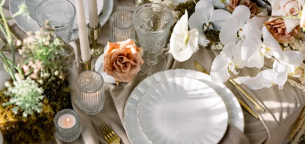A close-up of an elegant event table setting featuring white plates, gold cutlery, glass votives, and floral arrangements.