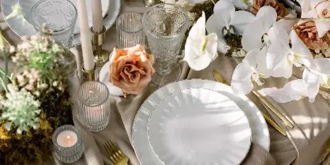 A close-up of an elegant event table setting featuring white plates, gold cutlery, glass votives, and floral arrangements.