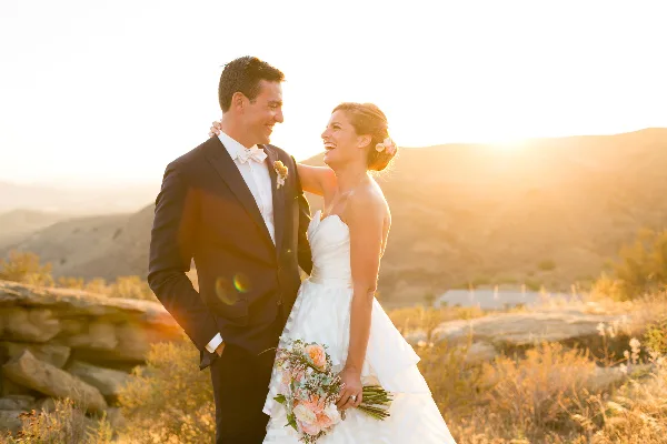 A bride and groom laughing together outdoors during golden hour, with the bride holding a bouquet of pink flowers.