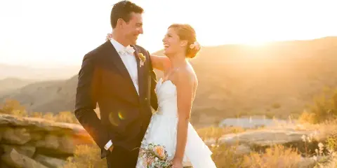 A bride and groom laughing together outdoors during golden hour, with the bride holding a bouquet of pink flowers.