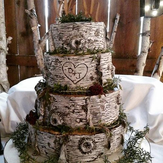 A four-tiered wedding cake decorated with a realistic birch bark pattern, greenery, and a heart monogram.