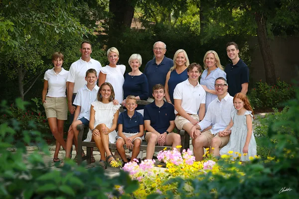 A professional outdoor family portrait of a multi-generational group posing on a bench in a garden setting.