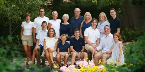 A professional outdoor family portrait of a multi-generational group posing on a bench in a garden setting.