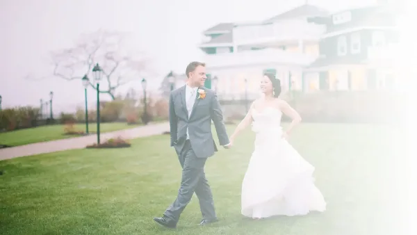 A bride and groom walking hand-in-hand on a grassy lawn in front of a large house.